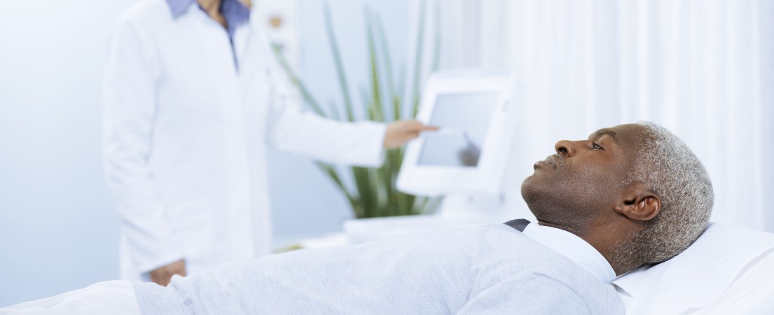 Foto de um paciente negro deitado na maca ao lado de uma médica branca que, de pé, ao lado da maca, toca na tela de um monitor