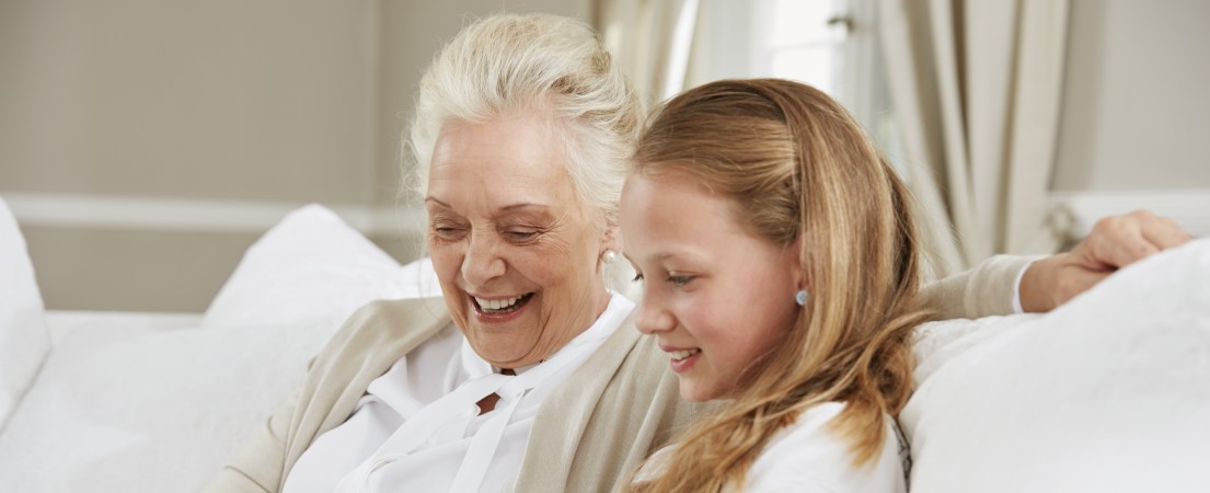 Foto de uma idosa branca sentada no sofá ao lado de uma garota. As duas estão olhando para baixo, sorrindo, ao que parece ser uma tela.