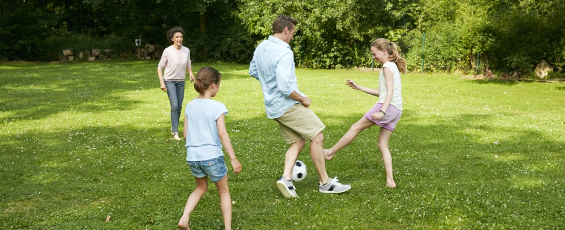 Na foto, um homem e uma mulher brancos jogam futebol com duas crianças brancas em uma área gramada e arborizada.