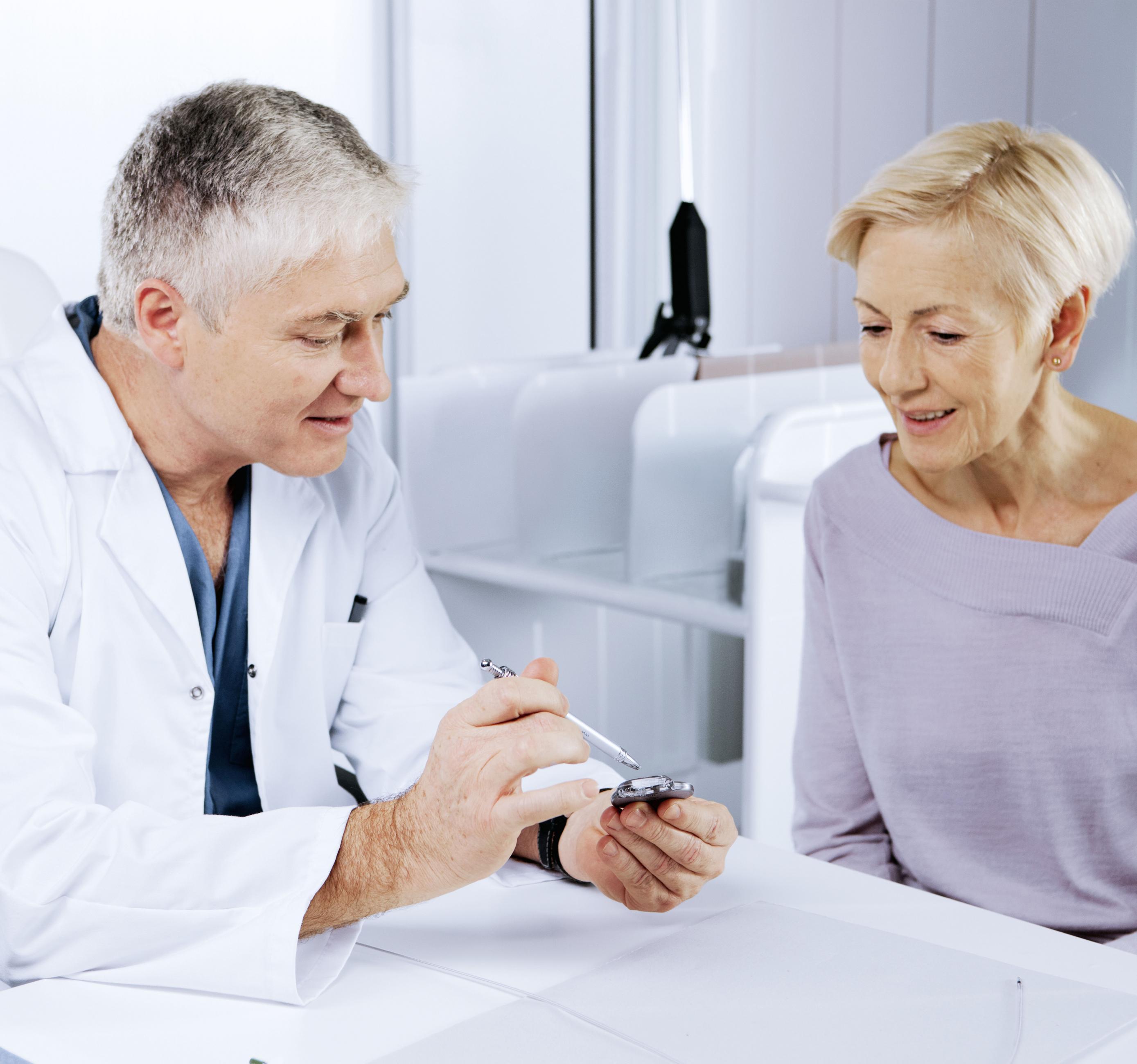 Na imagem, há um médico e uma mulher sentados próximos, em um consultório. Ele está mostrando um dispositivo para ela, que observa atentamente.