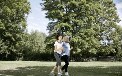 Na foto, duas mulheres brancas de meia-idade correm em uma área de grama e árvores