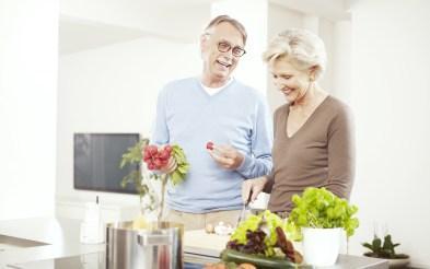 Na foto, um casal heterossexual de meia-idade está sorrindo em torno de um balcão na cozinha. O homem segura um ramo de rabanetes e a mulher está com uma faca cortando algo em cima de uma tábua. No balcão também estão folhas verdes e roxas e uma panela