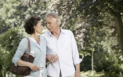 Na foto, em um ambiente arborizado, a imagem é de um homem e uma mulher de meia-idade, brancos, se olhando nos olhos de mãos dadas na altura da cintura