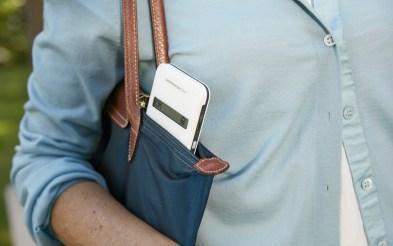 Na foto, o tronco de uma mulher branca com uma bolsa a tiracolo onde sobressai um aparelho branco semelhante a um smartphone 