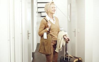  Foto de uma mulher branca de meia-idade com uma bolsa a tiracolo e segurando a alça de mala. Ela tem um casaco pendurado no braço e está em um corredor claro com várias portas e uma escada.