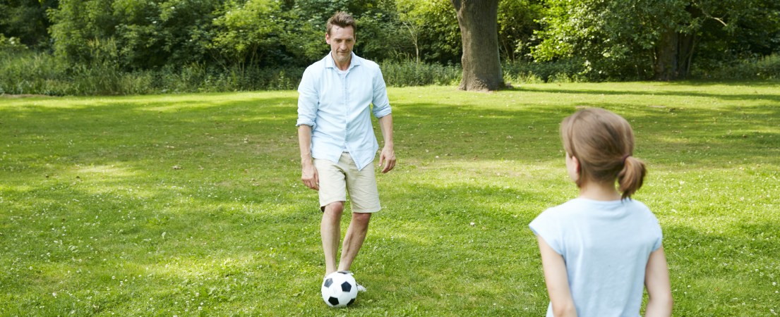 Foto de um homem brincando de bola com uma criança em uma área de grama e árvores.