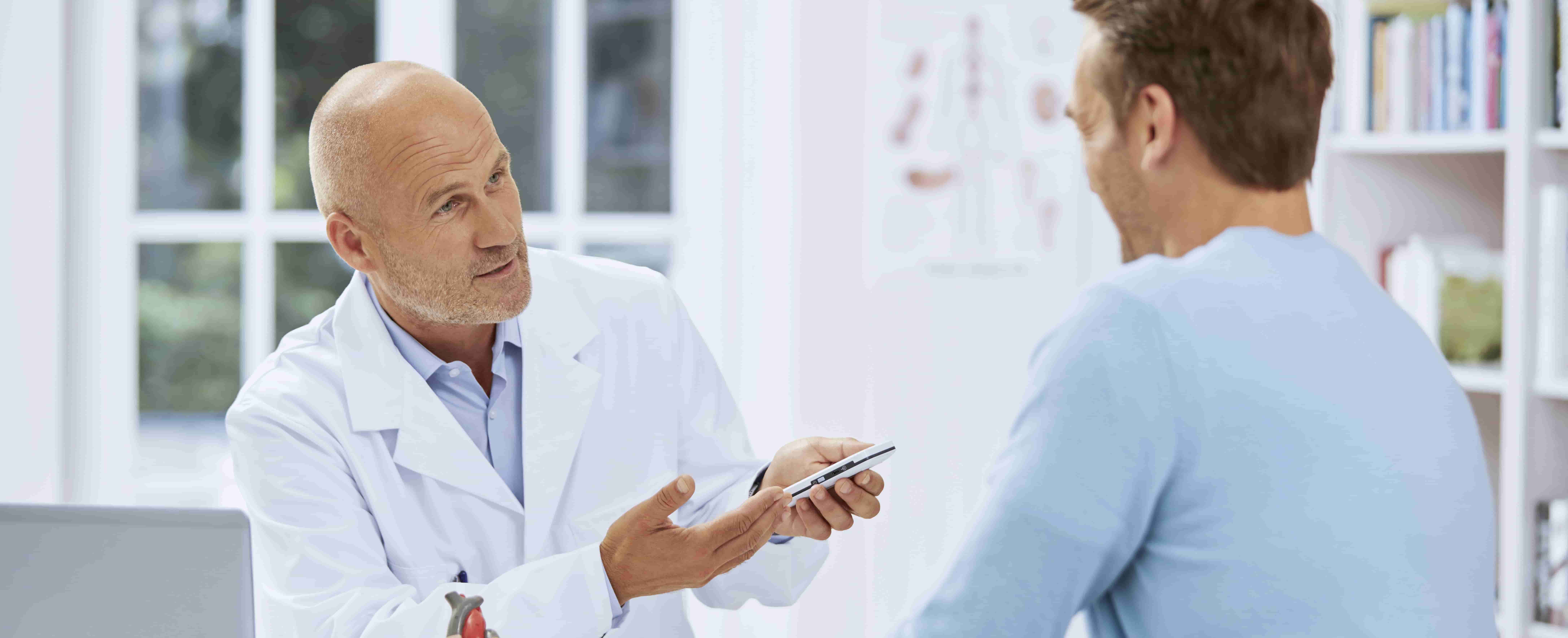 Foto de um médico em primeiro plano, branco, em ambiente hospitalar, mostrando a um homem branco, de perfil, um dispositivo semelhante a um smartphone