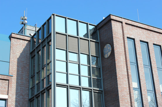  Foto de cima para baixo de parte de um prédio de fachada marrom com espelhos. Há duas antenas no topo e, em uma das colunas, próximo ao topo, há um círculo prateado com as iniciais BIO