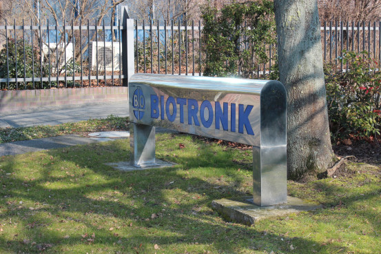 Na foto, uma placa prateada em posição horizontal onde estão grifados em azul o logotipo e nome BIOTRONIK. A placa está fixada próxima a uma árvore e instalada em uma área gramada. . 