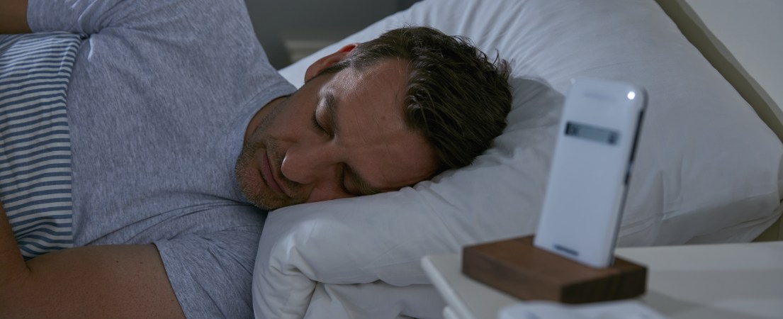 Foto de um homem branco dormindo em uma cama com a cabeça em um travesseiro e, ao lado, o dispositivo CardioMessenger apoiado em suporte sobre uma mesa de cabeceira