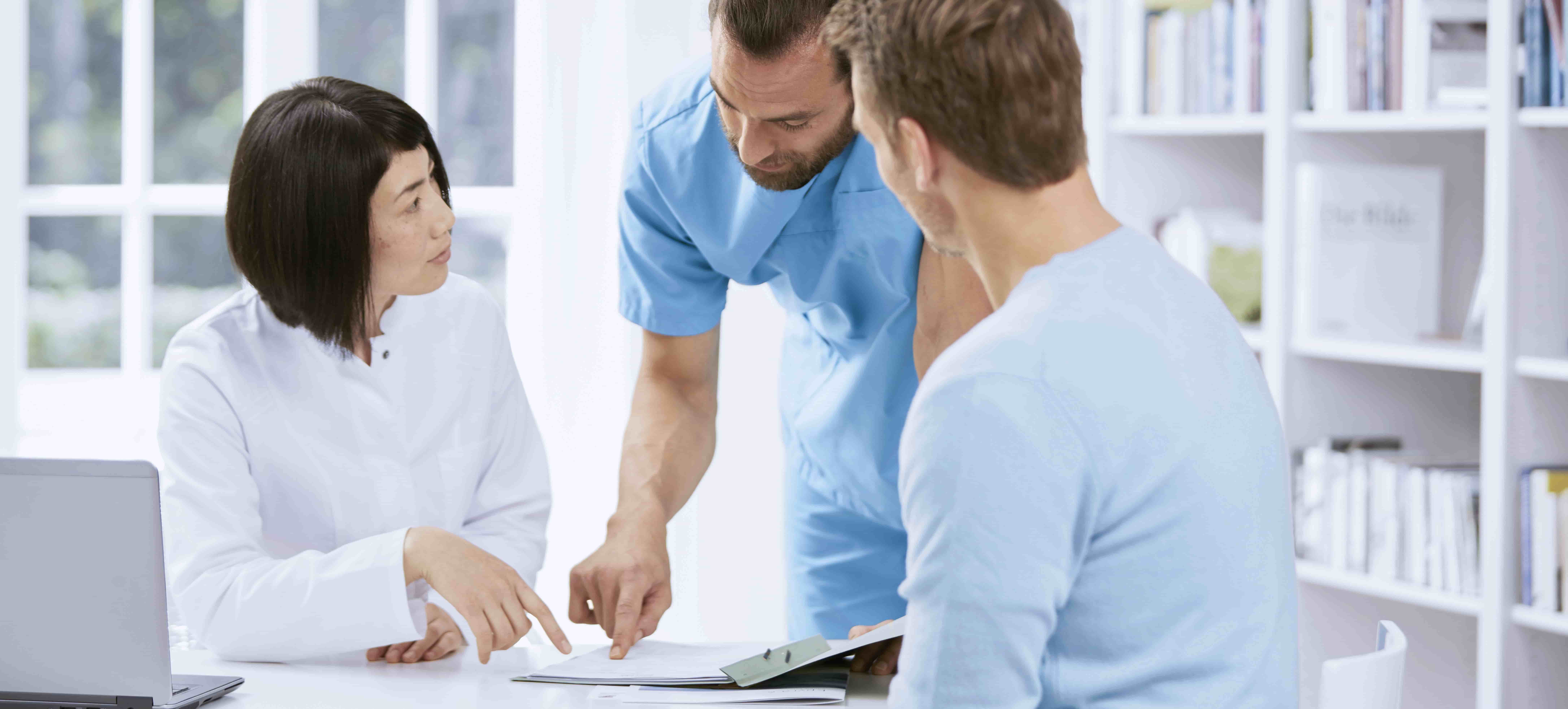 Foto de dois profissionais de saúde, um homem branco e uma mulher asiática, em ambiente hospitalar, de frente para um homem branco sentado, analisando um documento sobre a mesa