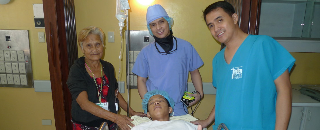 Na foto, de frente, dois profissionais de saúde e uma mulher de meia-idade em volta de um paciente jovem. Ele está deitado em uma maca, de touca cirúrgica, ao lado de um suporte de metal em que está pendurado uma bolsa de soro