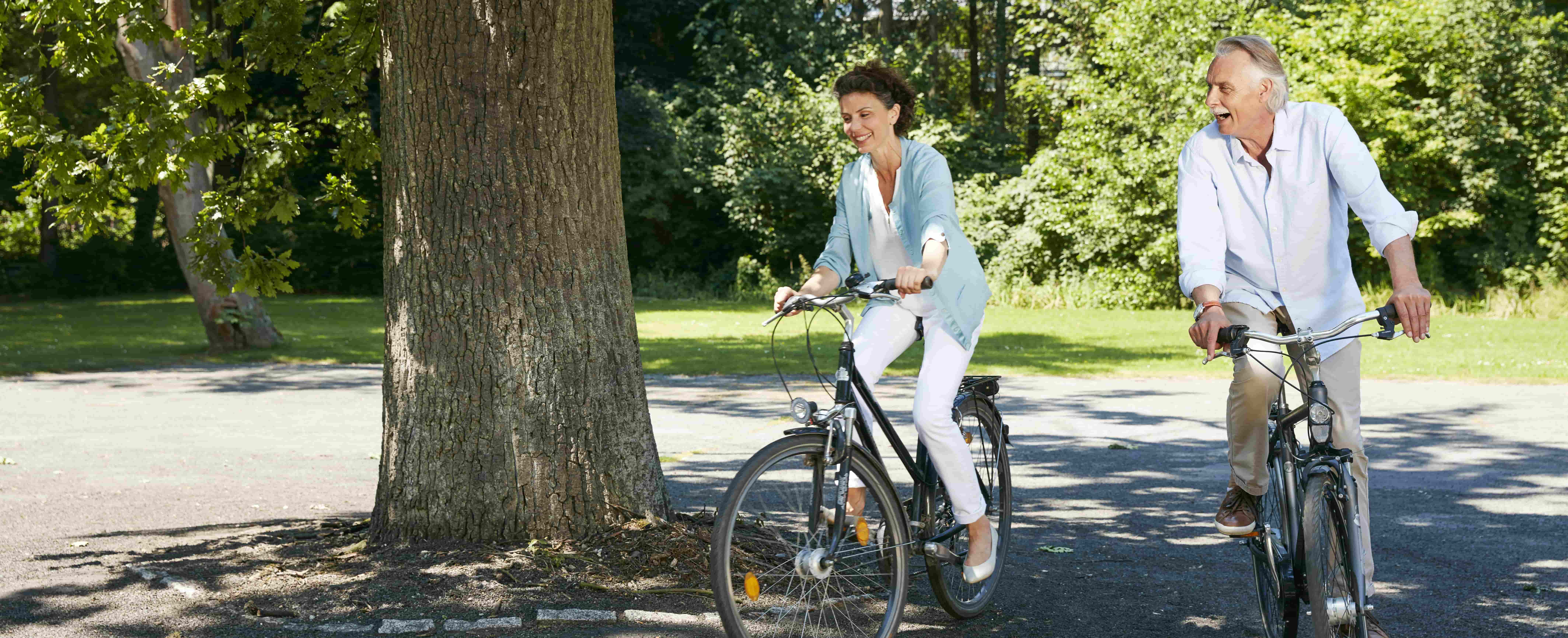 Na foto, há um homem e uma mulher em uma área arborizada. Eles estão andando de bicicleta.
