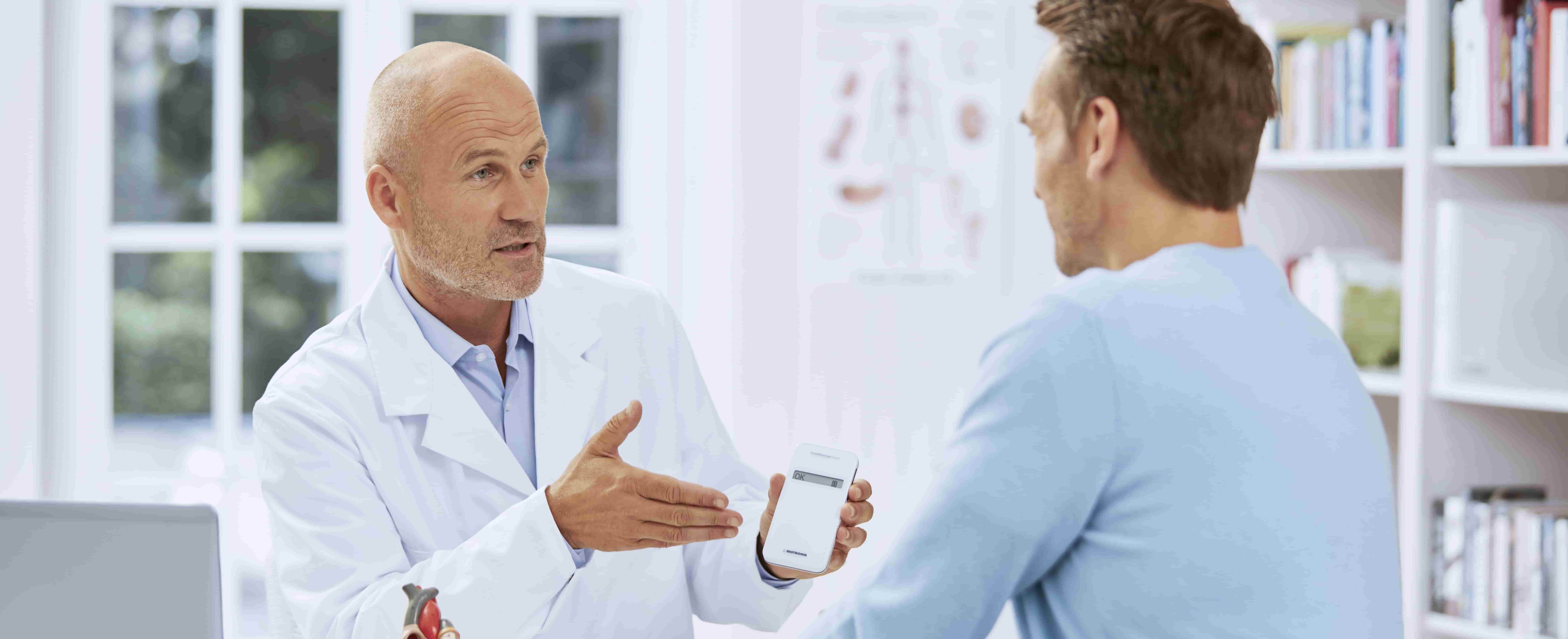 Foto de um médico branco em primeiro plano, sentado em ambiente hospitalar, mostrando a um homem branco, de perfil, um aparelho branco do tamanho de um pequeno smartphone