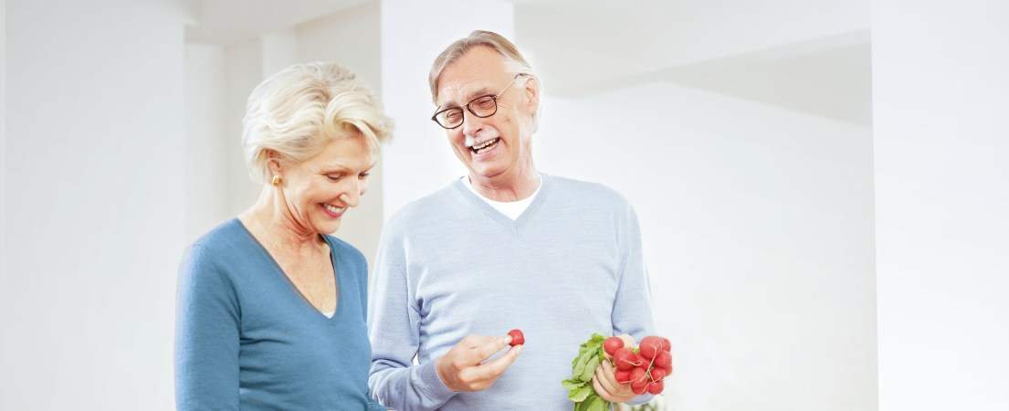 Na foto, um homem e uma mulher de meia-idade estão em um ambiente de paredes claras. O homem, olhando para a mulher sorrindo, segura um ramo de rabanetes. 