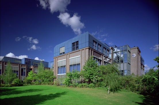 Na foto, a partir de um ambiente externo ensolarado, os fundos de um prédio de fachada marrom e azul em uma área com grama verde e algumas árvores de pequeno porte. O céu é azul com poucas nuvens 