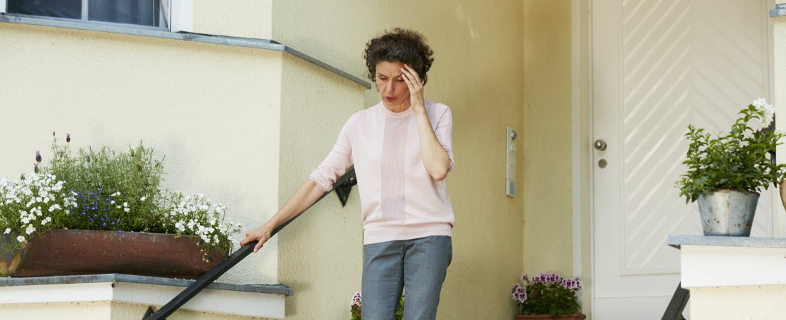 Foto de uma mulher branca, de meia idade, descendo as escadas com uma mão no corrimão e outra  na têmpora, expressando desconforto.