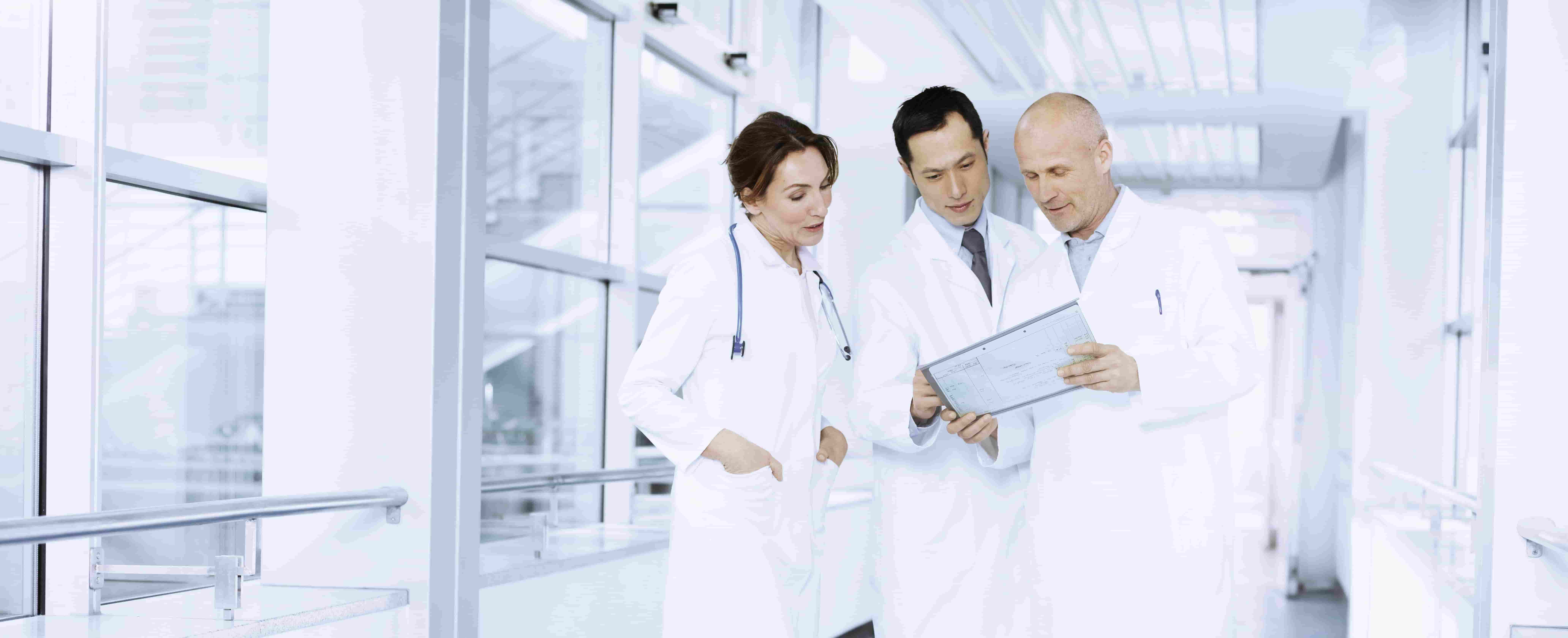 A foto mostra três médicos: dois homens e uma mulher. Ambos estão parados no corredor de um hospital, olhando para um papel e analisando dados. 