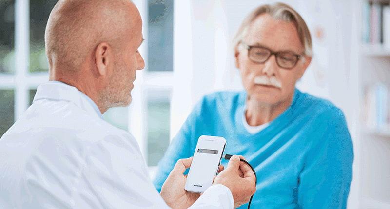 Fotografia com um médico branco segurando um dispositivo e explicando para um paciente branco, de aproximadamente 65 anos, como funciona o sistema BIOTRONIK Home Monitoring® 
