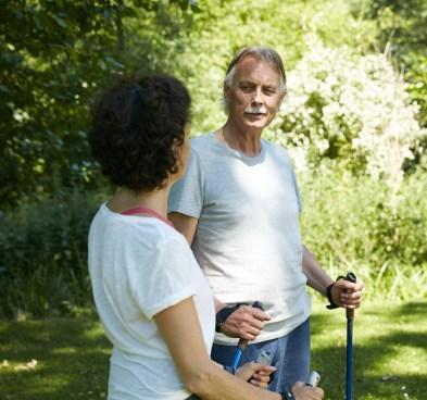 Foto de um homem e uma mulher brancos em um parque, se olhando, na pausa de um exercício físico em que seguram dois bastões, um em cada mão 