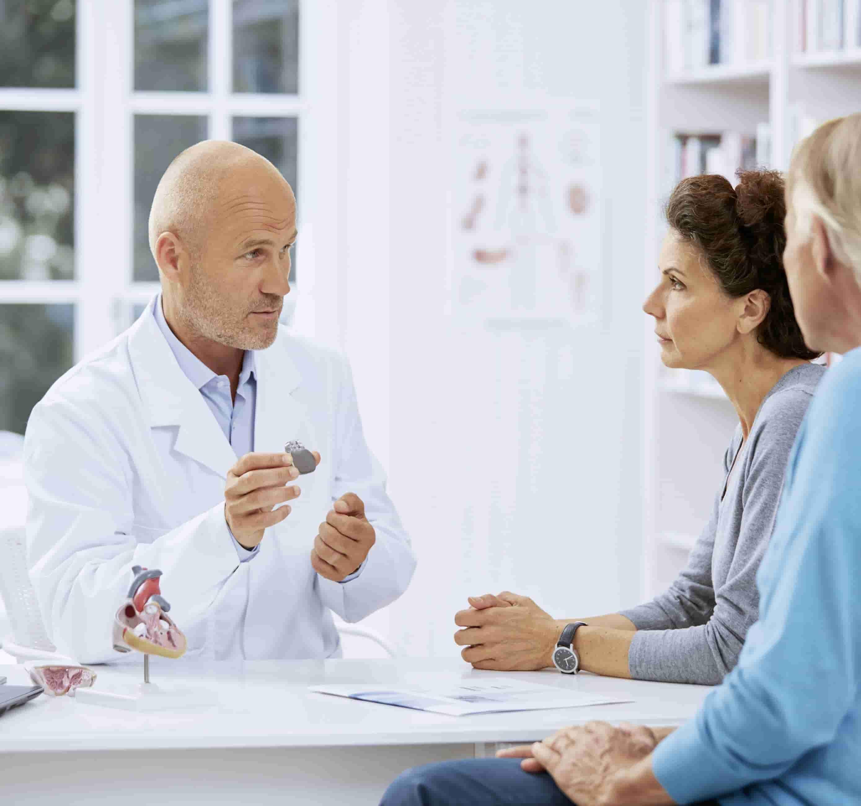 Foto de um médico em primeiro plano, branco, em ambiente hospitalar, mostrando a uma mulher branca de meia-idade, de perfil, um pequeno aparelho de cor cinza. Ao lado da mulher está de perfil um homem branco sentado