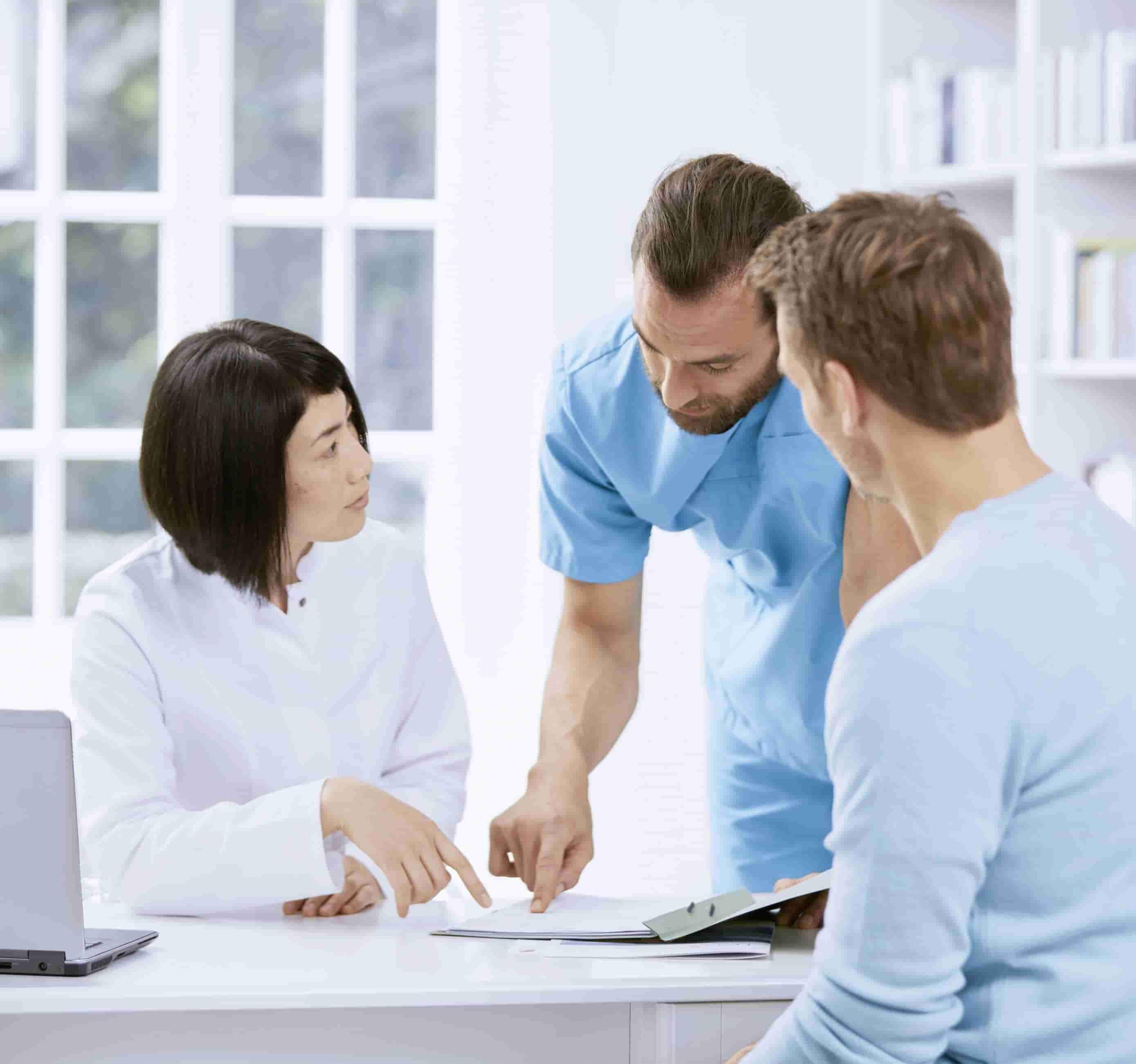 Foto de dois profissionais de saúde, um homem branco e uma mulher asiática, em ambiente hospitalar, de frente para um homem branco sentado, analisando um documento sobre a mesa