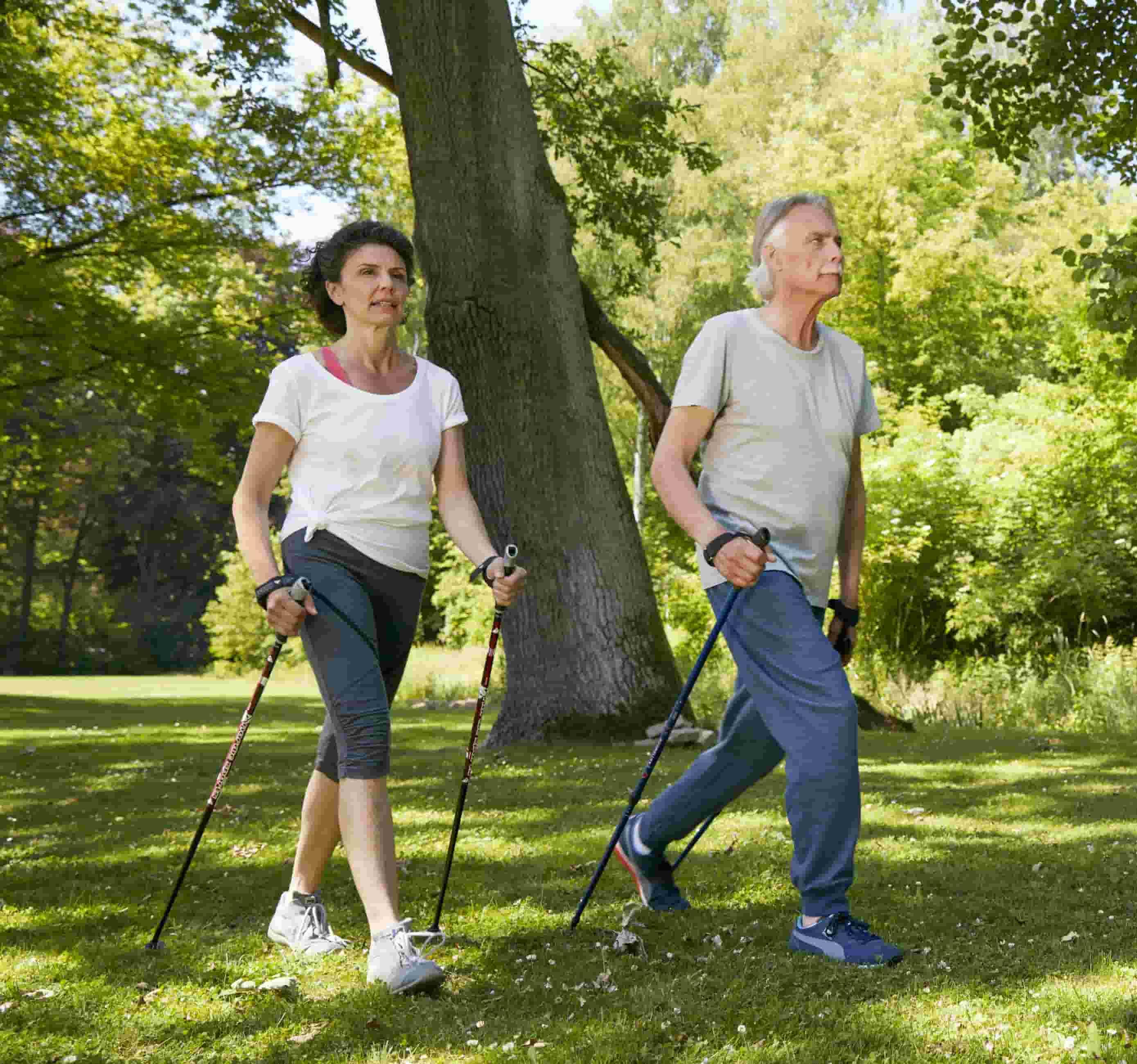 Foto de casal de meia-idade no parque segurando cada um dois bastões para facilitar a caminhada 