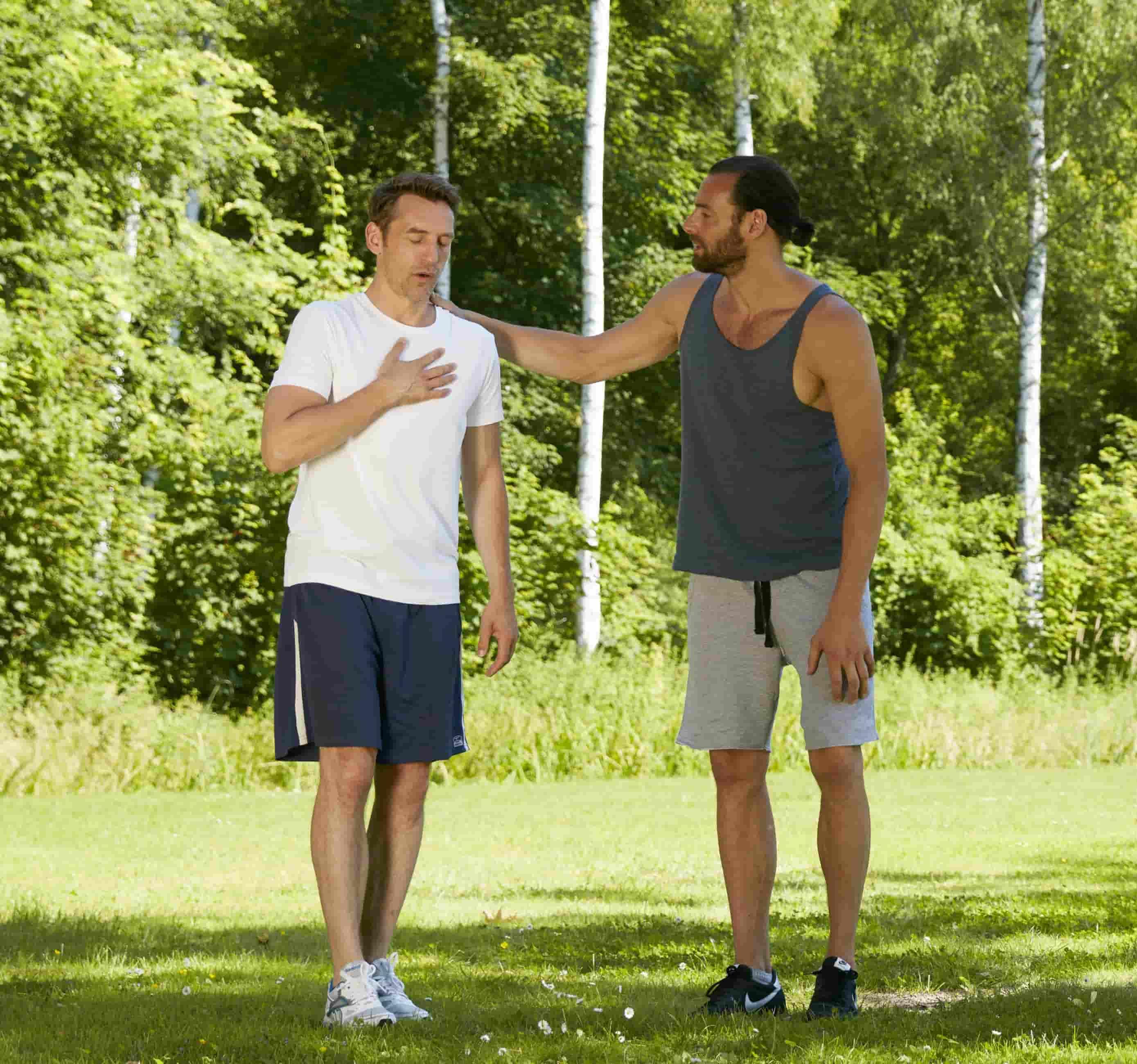 Na foto, há dois homens com roupas de atividade física em um parque. Um deles, a esquerda, está com a mão no peito, demostrando estar passando mal. O outro está com a mão em seu ombro, amparando-o.