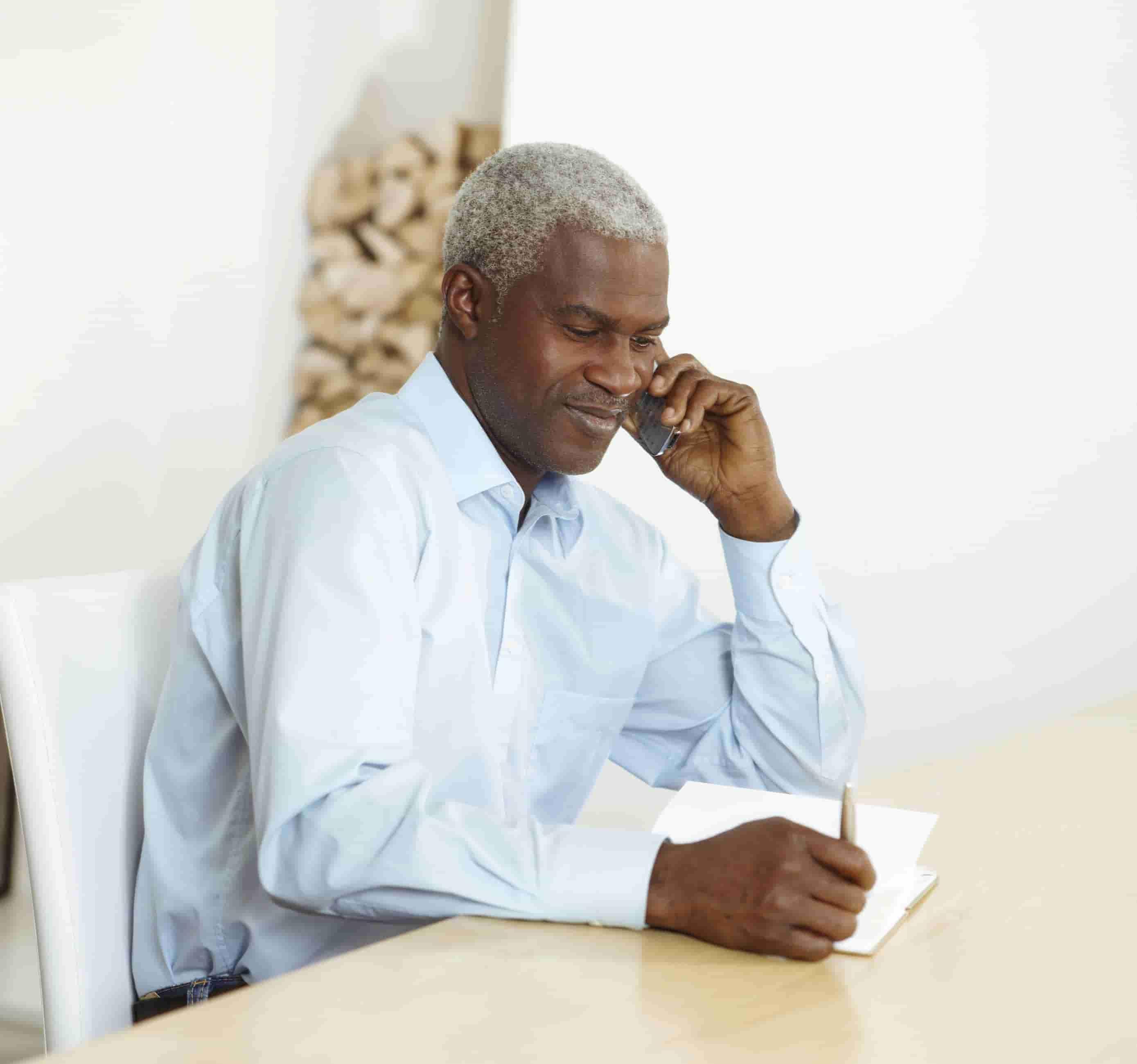 Foto de homem negro grisalho sentado à mesa e falando ao telefone enquanto segura uma caneta sobre um bloco de anotações. 