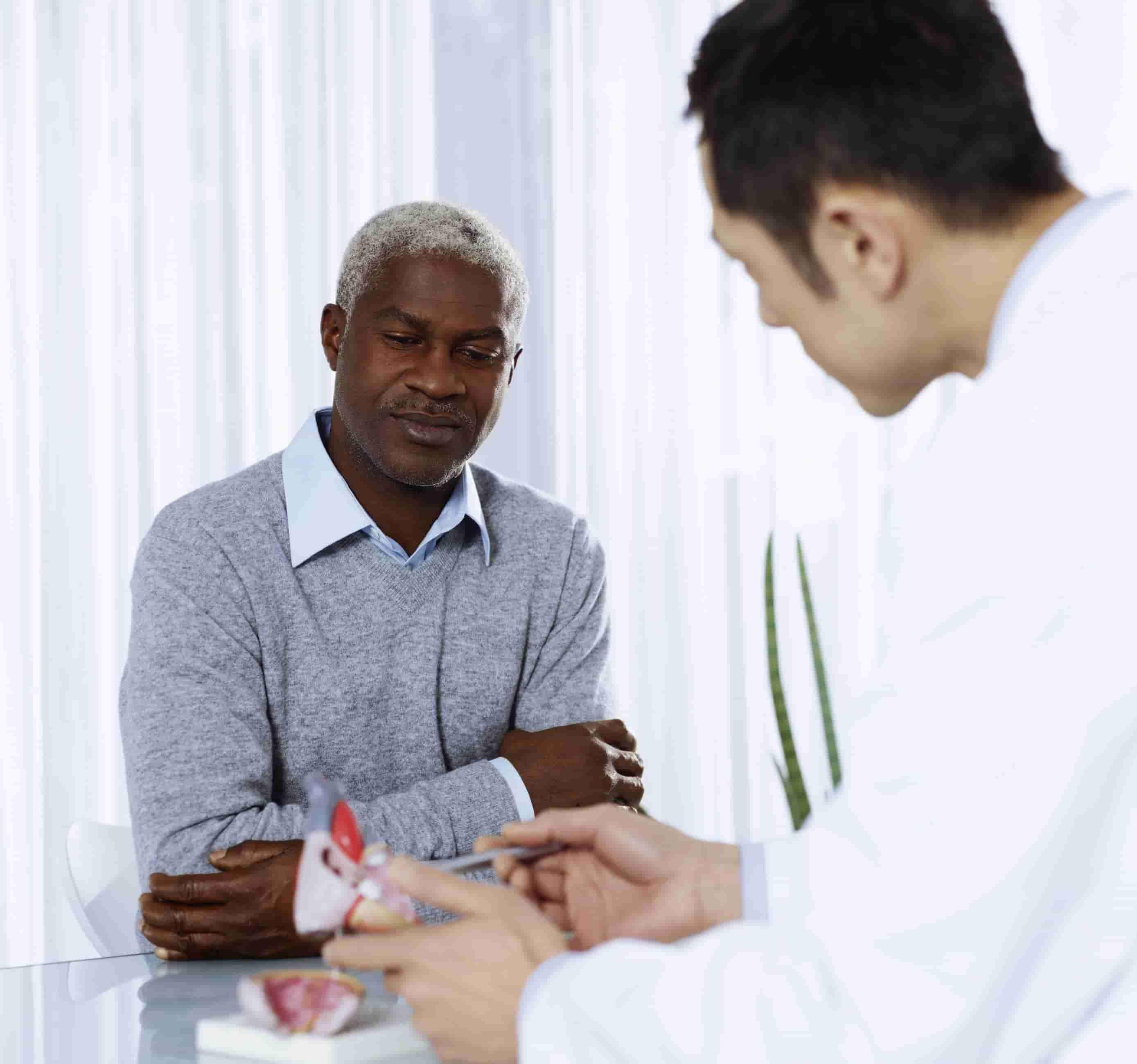 Na foto, há um senhor negro de meia idade sentado a frente de um médico que está lhe explicando algo, com uma maquete de coração na mão.