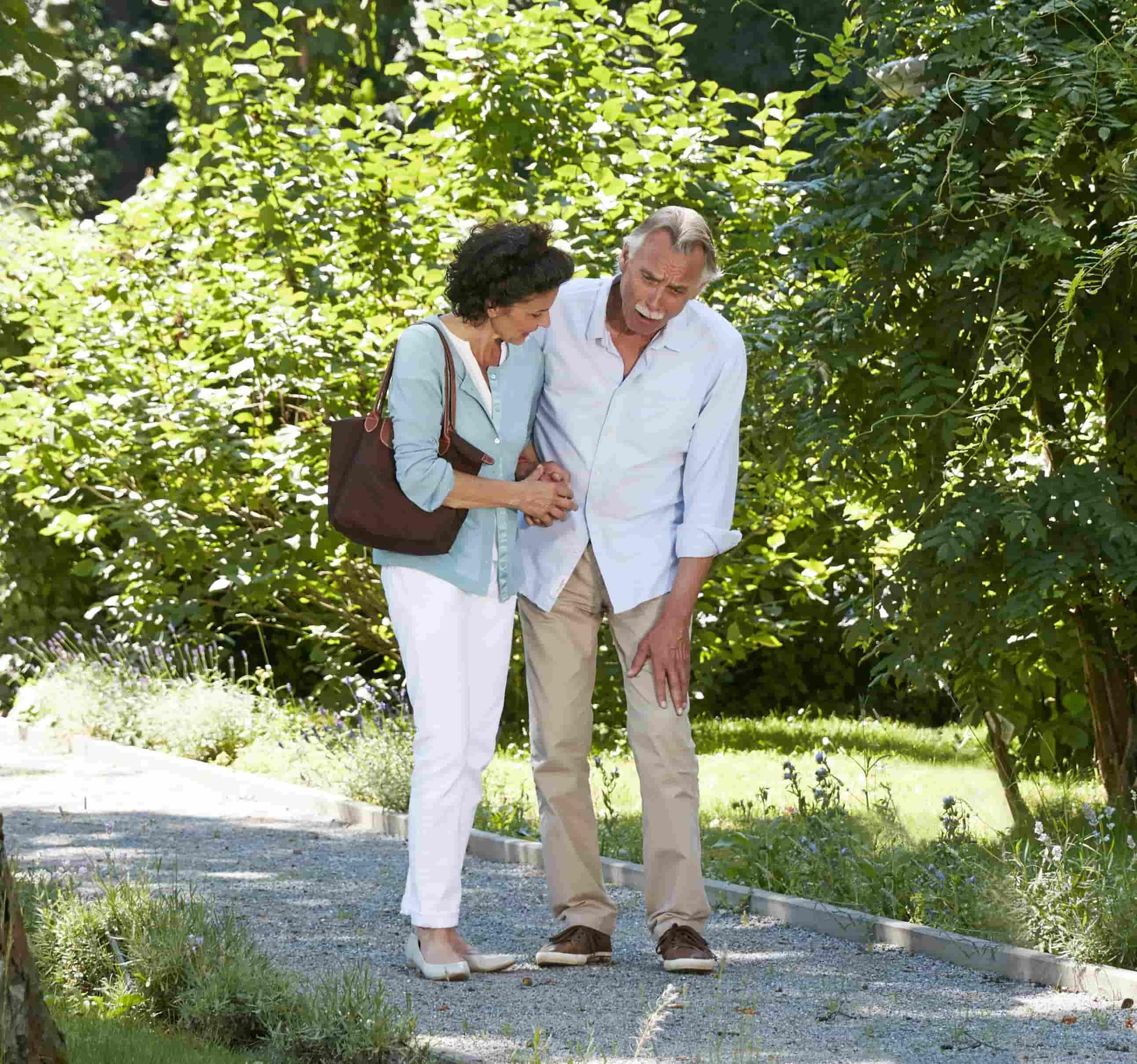 A imagem mostra um homem e uma mulher andando em um parque. O homem, à direita, demonstra estar sentindo dor, enquanto a mulher, à esquerda, o ampara.