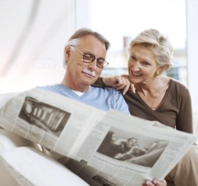 Na imagem, há uma homem e uma mulher, idosos. Ambos estão sentados olhando para um jornal aberto, que está na mão do senhor.