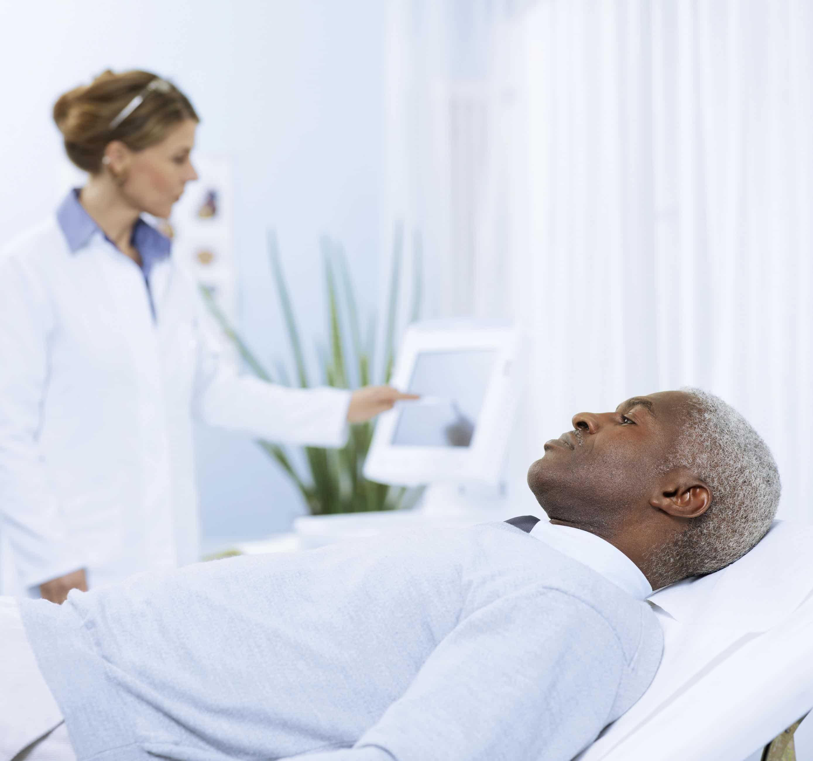 Foto de um paciente negro deitado em uma maca e sendo atendido por uma médica branca que mexe em um monitor. 